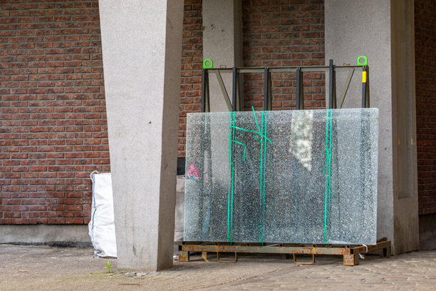 破裂的玻璃板用运输带固定，垂直放置在一个托盘上，位于混凝土棚下，背景是砖墙。照片展示了在建筑工地上为安装或运输准备的建筑材料。图片下载
