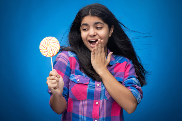 可爱的年轻女孩，直发，面带微笑，手持圆形棒棒糖，背景为蓝色。图片下载