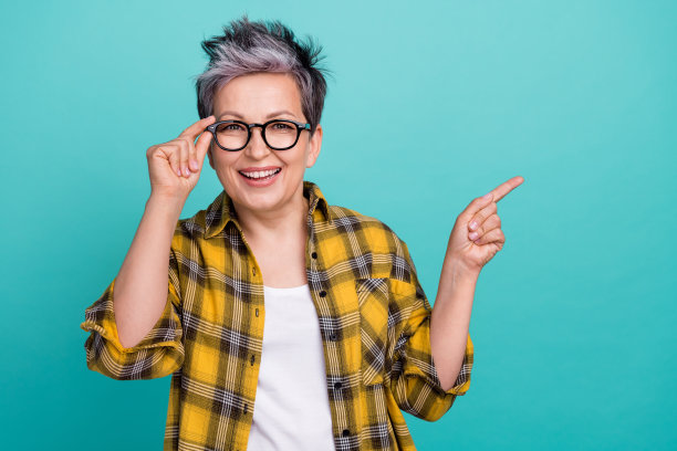 迷人的祖母在青色背景下穿着时尚的黄色格子服装，触摸眼镜，指向空白处的照片肖像。图片下载