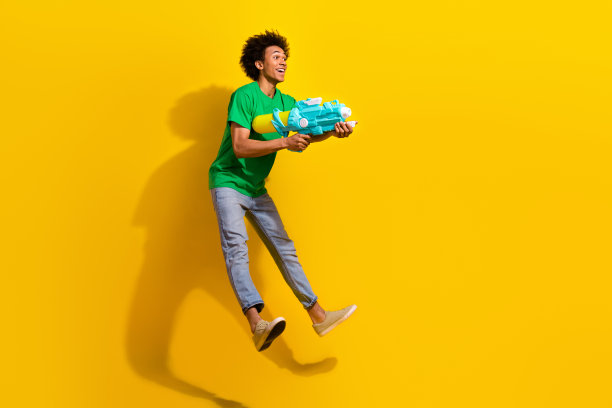 全身照片，吸引人的搞笑男士穿着绿色T恤，跳得很高，享受水上游戏，背景为空旷的黄色。图片下载