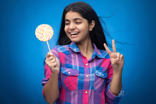 可爱的年轻女孩，直发，手持圆形棒棒糖，面带微笑，背景为蓝色。图片下载