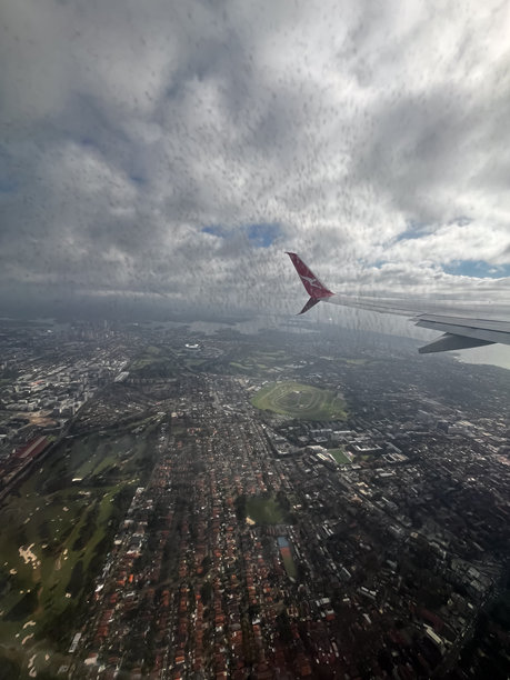 一张情绪化的悉尼郊区航拍照片，通过雨点斑驳的飞机窗户拍摄，画面中有一架在厚厚云层下的澳航飞机机翼。图片下载