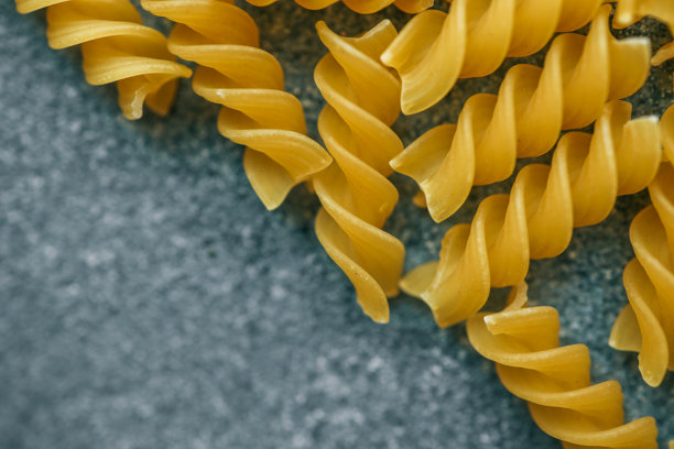未煮的黄色螺旋面条的特写照片，展示了螺旋形状和干燥的质地，背景为纹理灰色石材，呈现简约的食物构图。图片下载