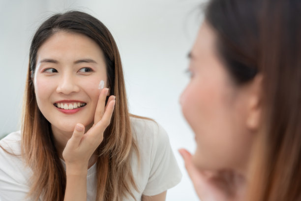 美丽的亚洲女性微笑着使用护肤霜。健康女性的面孔涂抹护肤霜和化妆品。护肤霜广告，抗皱，婴儿般的肌肤，美白，保湿，收缩毛孔精华。图片下载