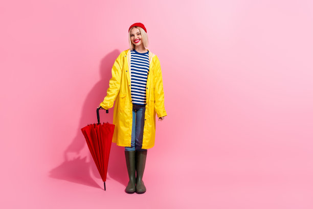 穿着雨衣的快乐漂亮女人手持阳伞在粉色背景的空旷空间中行走的全身照片图片下载