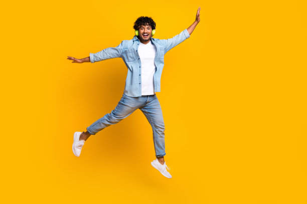 穿着牛仔夹克的搞笑兴奋男孩在空旷的黄色背景下高高跳起，听着音乐的全身照片图片下载