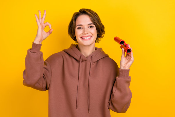 年轻乐观的女孩，短发，探索旅游，做出“好”的手势，手持望远镜，放大图片，穿着毛衣，背景为黄色。图片下载