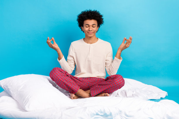全身照片，平静积极的人赤脚坐在舒适的床单上冥想，背景为蓝色。图片下载