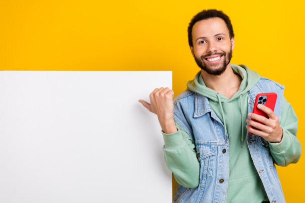 年轻男子的照片肖像，手指指向白色广告牌，手持设备，穿着时尚的卡其色衣服，背景为黄色。图片下载