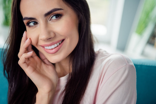 特写裁剪照片，展现可爱积极的女孩灿烂的微笑，手臂轻触颧骨，享受室内的闲暇时光。图片下载