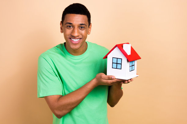 时尚绿色服装的英俊年轻男性手持小房子抵押贷款的照片肖像，背景为米色。图片下载