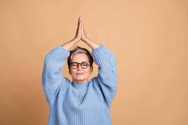 可爱的退休女性在米色背景下穿着时尚的蓝色针织服装，双手举起祈祷姿势的照片肖像图片下载