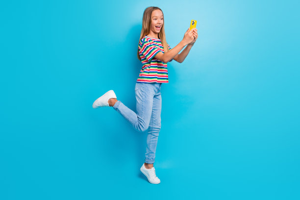 迷人的少女穿着彩色T恤和牛仔裤，在蓝色背景下用智能手机自拍的全身照片图片下载