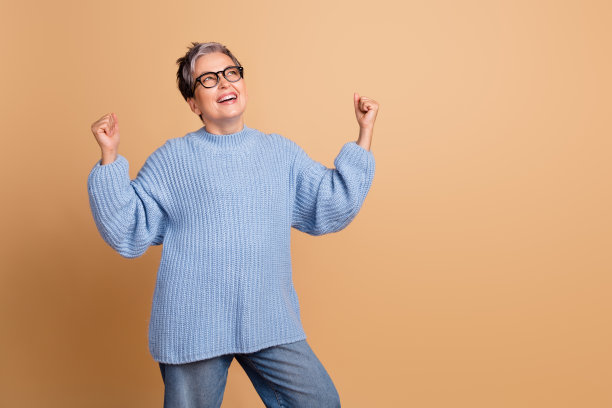 可爱的退休女性在米色背景下穿着时尚的蓝色针织服装，举起双拳庆祝的照片肖像图片下载