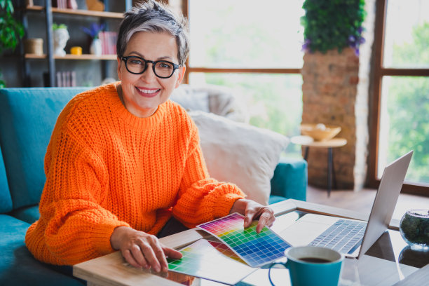 穿着橙色毛衣的快乐积极老年女士在公寓室内手持色彩调色板和现代设备的照片图片下载