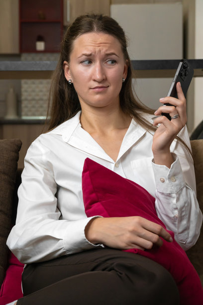 年轻女性，长棕色头发，穿着白衬衫，坐在沙发上，手持智能手机，面露困惑，惊讶于她所阅读的令人不安的消息。竖版照片图片下载