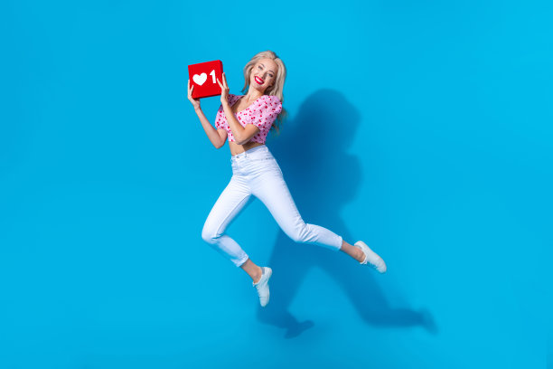 穿着时尚樱桃印花服装的可爱年轻女士在蓝色背景上跳跃，手中握着心形图标的全身照片图片下载