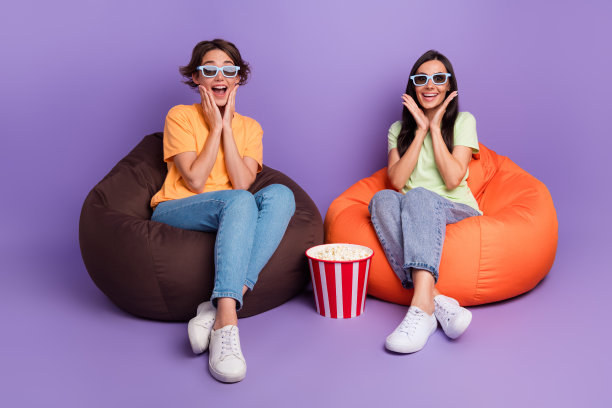 两位好朋友的全身照片，女孩们手托着脸颊，观看新的007特工电影，3D影院，背景为紫色。图片下载