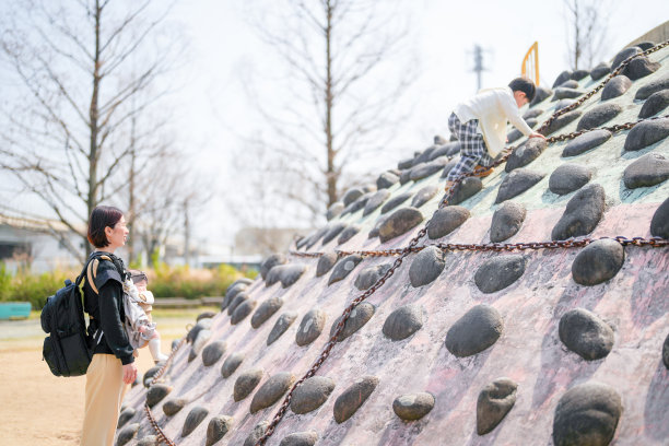 在福冈的公园里，春天，一个3岁的男孩正在攀爬一个岩石结构。他的妈妈和6个月大的妹妹在一旁观看。这张照片捕捉了一个温馨而紧密的家庭时刻，背景是宁静的户外环境。图片下载