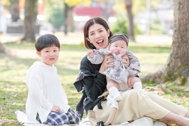 在福冈的公园春天，一位三十多岁的妈妈与她三岁的儿子和六个月大的女婴坐在一起，面带微笑。这张照片展现了一个宁静而难忘的家庭时刻，融入大自然中。图片下载