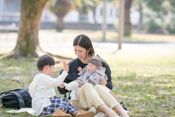 在福冈的公园春天，一位三十多岁的妈妈与她三岁的儿子和六个月大的女婴坐在一起，笑容满面。这张照片展现了春季环境中一个宁静而温馨的家庭时刻。图片下载