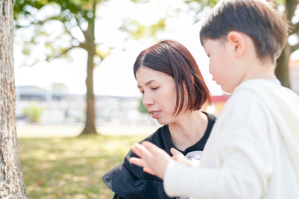 在福冈的公园春天里，一位三十多岁的妈妈与她三岁的儿子和六个月大的女婴坐在一起，笑容满面。这张照片展现了春季环境中一个宁静而温馨的家庭时刻。图片下载