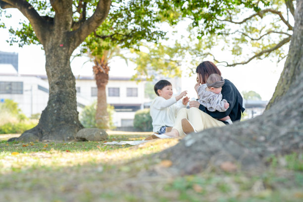 在福冈的公园里，春天，一位日本妈妈抱着她六个月大的女婴。她三岁的儿子坐着吃零食。这张照片捕捉了一个宁静而充满爱的时刻，展现在一个安静的户外环境中。图片下载