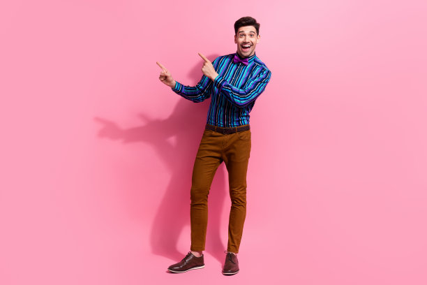 穿着复古衬衫和蝴蝶结的搞笑年轻男子在粉色背景上指向空白空间，拍摄的全身照片，适合精品广告。图片下载