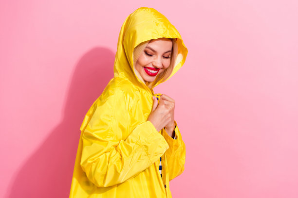 年轻漂亮的女士在粉色背景上藏着一件保护性的黄色防水秋季外套，作为商店广告的个人资料照片。图片下载