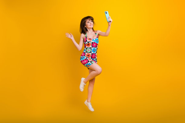 穿着花卉印花服装的无忧无虑、兴奋的女性在黄色背景下高高跳起，录制视频博客的全身照片图片下载