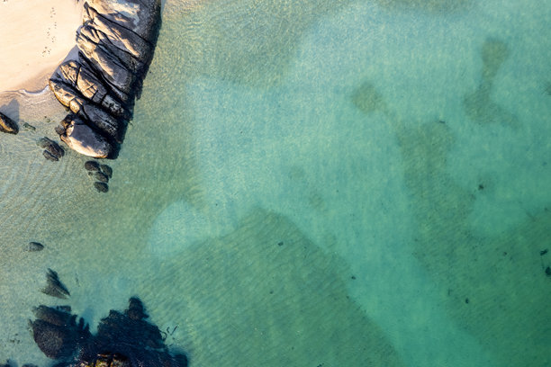 从上方俯瞰清澈的水、沙滩和黑色岩石。俯视航拍照片。夏季背景。图片下载