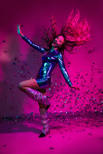 在霓虹粉色背景下，一位穿着闪亮蓝色连衣裙和光亮靴子的时尚女性正在进行生动的庆祝活动，竖版照片。图片下载