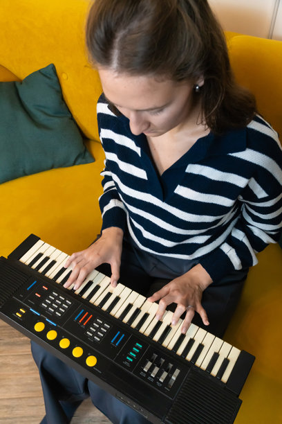 年轻女性坐在舒适的黄色沙发上，专注于弹奏电子琴，在温馨的家居环境中创作音乐。竖版照片图片下载