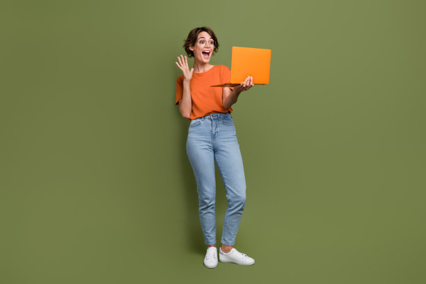 穿着橙色T恤的搞笑女孩在绿色背景下，戴着眼镜，向笔记本电脑挥手，进行视频通话的全身照片。图片下载