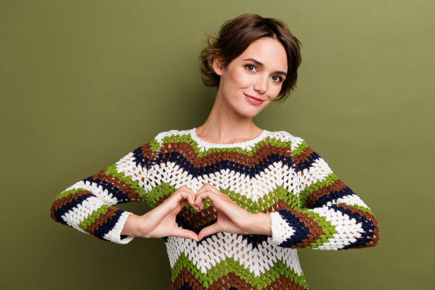 迷人的年轻女性在卡其色背景下展示心形手势，穿着时尚印花针织服装的照片肖像。图片下载