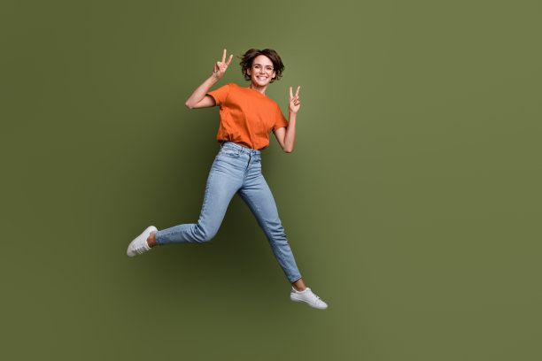 穿着时尚衣服、佩戴眼镜的满意女孩在绿色背景上跳跃，做出V字手势的全身照片图片下载