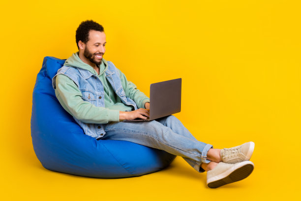 拉丁裔自由职业者商人在黄色背景上坐在坐垫上使用笔记本电脑的全身照片，专注于社交媒体营销目标。图片下载