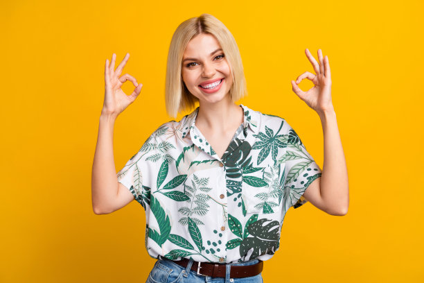 漂亮年轻女孩的照片肖像，展示双手“OK”手势，穿着时尚的叶子印花服装，背景为黄色。图片下载