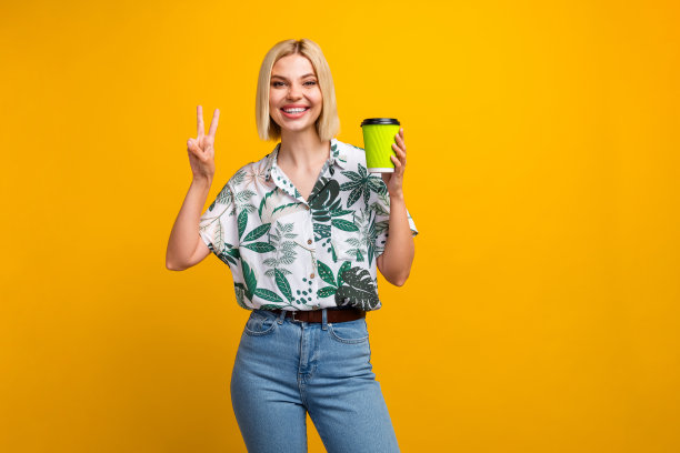 漂亮年轻女孩的照片肖像，做出V字手势，手持纸杯，穿着时尚的叶子印花服装，背景为黄色。图片下载