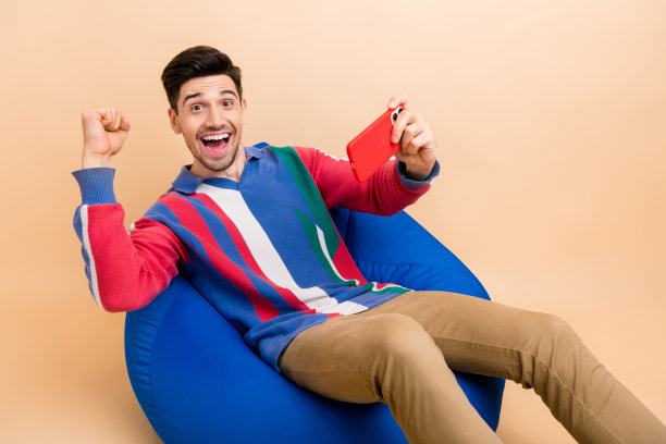 全身照片的男孩坐在豆袋椅上，握拳庆祝在智能手机应用中赢得他的第一场比赛，穿着时尚的衬衫，背景为米色。图片下载