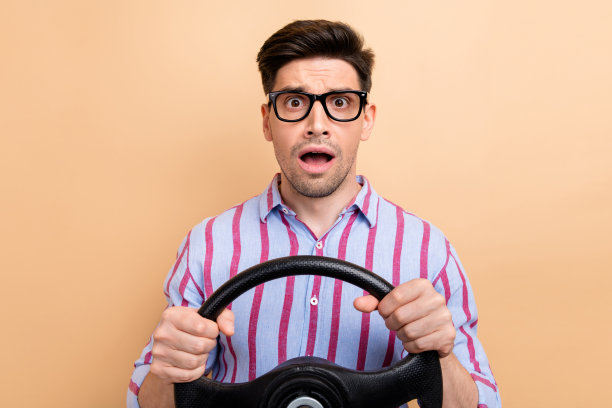 特写照片：年轻搞笑的困惑男子在学习如何驾驶，因观看汽车碰撞而感到压力，背景为米色。图片下载