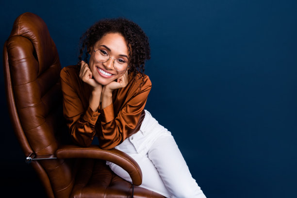 吸引人的年轻女性照片肖像，穿着时尚的棕色丝绸正装，坐在符合人体工程学的椅子上，背景为深蓝色。图片下载