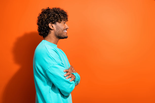 橙色背景上，微笑的印度男士侧面照片，双手交叉，展现出积极的态度。图片下载