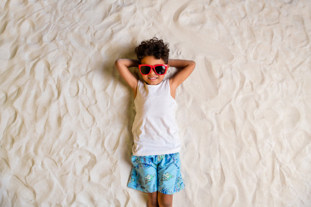 高角度拍摄的照片，阳光明媚的小男孩穿着白衬衫，躺在沙滩上，双手放在头后，享受海边的户外时光。图片下载