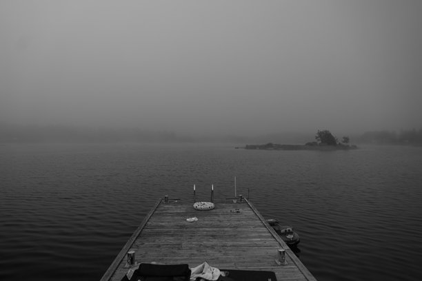 对称的单色照片，展示了一堵雾气笼罩的橡木码头，码头上散落着毛巾、玩具、泳衣和一张躺椅。浓雾遮住了瑞典斯德哥尔摩群岛格拉索岛的海湾尽头。图片下载