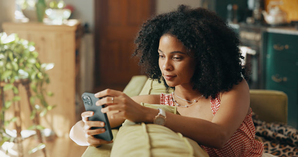 在家中沙发上放松的非洲女性，使用手机进行社交媒体、网络和沟通。公寓、客厅和正在使用智能手机进行在线聊天、互联网发布或周末网站浏览的人。图片下载