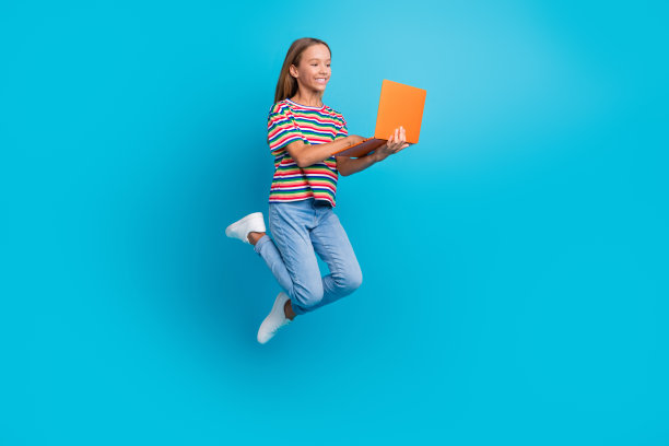 穿着条纹T恤的聪明少女在蓝色背景下跳跃，正在用笔记本电脑做作业的全身照片图片下载