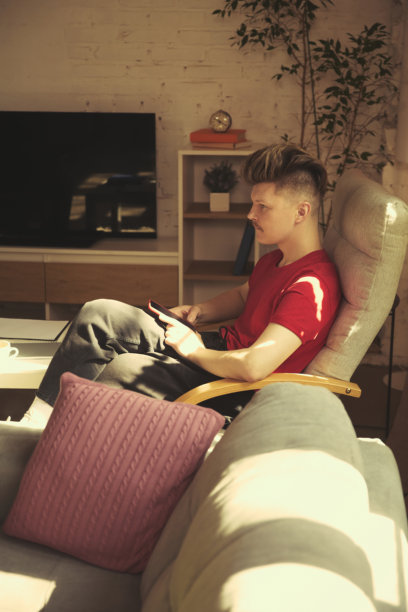 穿着红色衬衫的男子坐在时尚的扶手椅上，手持智能手机，注视着电视，现代化的客厅内饰。图片下载