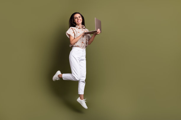 全身照片：一位快乐的女性自由职业内容经理在蹦床上跳跃，使用笔记本电脑，背景为卡其色。图片下载