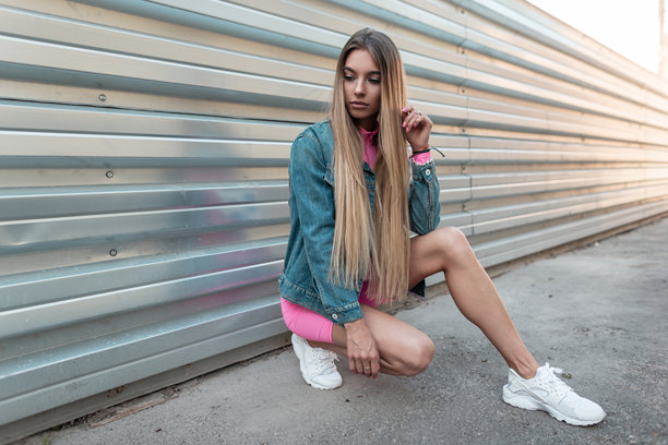 年轻漂亮的金发女子穿着时尚的夏季牛仔服和白色潮流运动鞋，坐在城市中一面金属墙旁，享受阳光明媚的夏日。现代女孩在户外休息。复古风格。图片下载